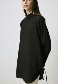 Zwarte langemouwen top met een hoge hals, zijstrikdetails en een comfortabele pasvorm, gemaakt van gladde stof en met een asymmetrische zoom.