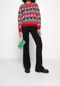 Rot-schwarzer gemusterter Pullover mit Schneeflocken und Rentieren; gerippte Bündchen und Saum. Schwarze Rippenhose und grüne Clutch-Tasche runden das Outfit ab.