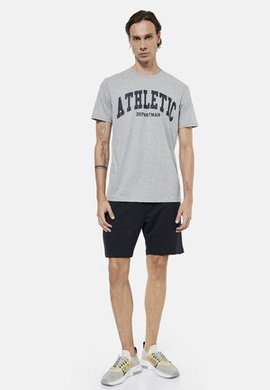 Mężczyzna stoi, ubrany w szarą koszulkę z nadrukiem "ATHLETIC", czarne szorty, białe i beżowe sneakersy, z tatuażami na ramionach na jednolitym tle.