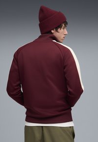 Mørkerød sweatshirt med høj krave, hvide sideløsninger og ribbede ærmer. Inkluderer en matchende mørkerød strikket hue.