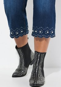 Botas negras hasta el tobillo con tachuelas metálicas y puntas afiladas, combinadas con jeans azul recortados que tienen dobladillos con forma de concha y adornos de perlas.