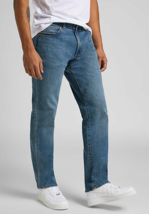 Jeans in denim blu con taglio dritto, caratterizzati da una vita media e delicate sfumature. Abbinati a sneakers bianche su uno sfondo grigio.