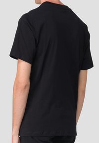 Uomo che indossa una semplice maglietta nera a maniche corte, visto di spalle su uno sfondo grigio chiaro semplice.