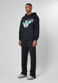 Felpa nera con design grafico che presenta un teschio, abbinata a pantaloni neri coordinati. Il modello indossa sneakers argentate. Tessuto morbido, vestibilità rilassata.