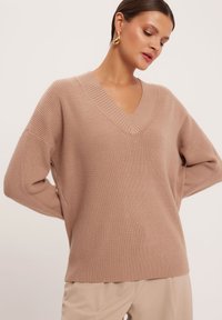 Beżowy sweter z dekoltem w serek, dziany z ribbowym wzorem, z obniżonymi ramionami i luźnym krojem. Minimalistyczny design, miękka tkanina i brak widocznych zapięć.