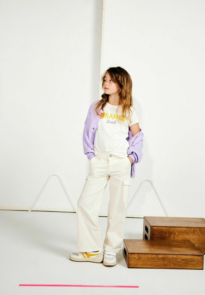 T-shirt blanche avec texte orange et jaune, cardigan lavande, pantalon cargo beige et baskets blanches avec accents jaunes. Fond uni.