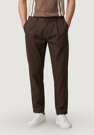 Uomo che indossa pantaloni plissettati marrone scuro e sneakers bianche, con le mani in tasca, su sfondo chiaro uniforme.