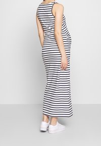 Frau steht seitlich und trägt ein ärmelloses Maxi-Kleid mit weißen und schwarzen Streifen sowie weiße Sneaker vor einem einfarbigen Hintergrund.