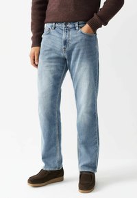 Gai zili džinsi ar taisnu kāju griezumu, piecām kabatām un kontrastējošu šuvju, valkāti ar tumši brūniem zamšādas apaviem.