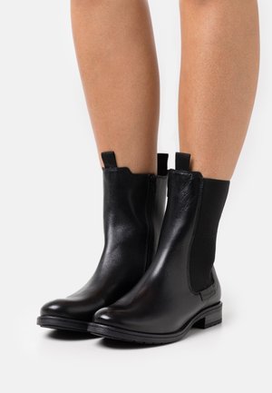 Bottes noires à la cheville en cuir brillant avec des panneaux élastiques sur les côtés, bout arrondi et petit talon empilé. Équipées de tirettes pour un enfilage facile.
