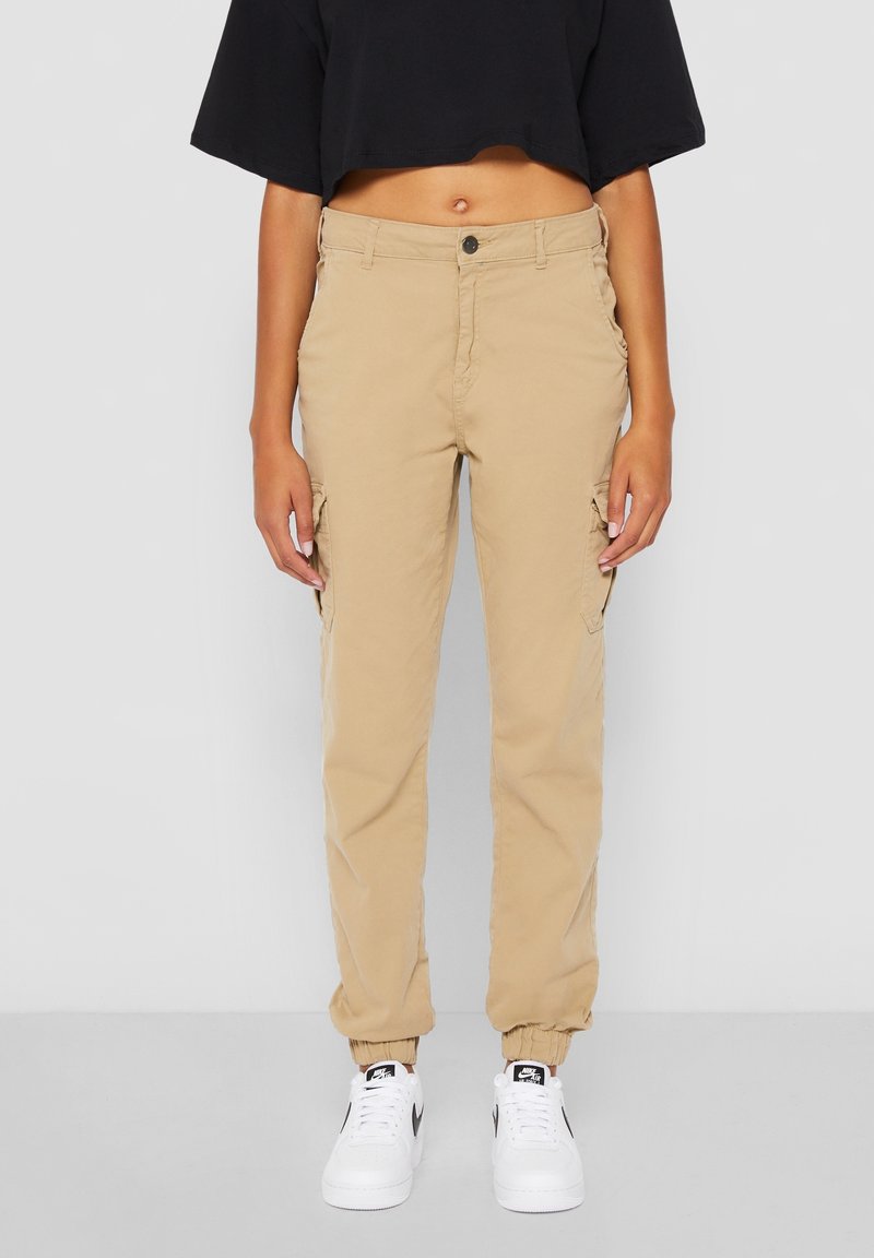 Personne portant un pantalon cargo beige avec des poignets élastiques, un t-shirt court noir et des baskets blanches Nike sur un fond uni.