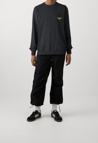 Sudadera negra con cuello redondo y logo amarillo, combinada con pantalones negros de corte holgado y zapatillas negras con detalles en blanco y suelas grises.