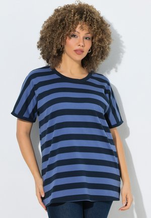 Vrouw met krullend haar die een losse blauw-zwarte horizontaal gestreepte korte mouw shirt draagt en tegen een lichte muur staat.