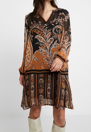 Kvinde iført knælængde kjole med lange ærmer og sort, brun og orange blomster- og stribet mønster, kombineret med lyse knæhøje støvler.