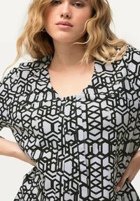 Schwarz-weißes Kleid mit geometrischem Muster, das über einen runden Ausschnitt, kurze Ärmel und Faltendetails verfügt. Hergestellt aus einem leichten Stoff.