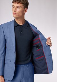 Hellblauer Anzug aus strukturiertem Stoff. Das Innere ist mit marineblauen Futter und roten Akzenten versehen und verfügt über mehrere Taschen. Darunter ein schwarzes Polohemd.
