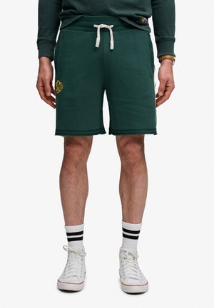 Mann trägt dunkelgrüne Shorts mit Kordelzug, weiße High-Top-Sneaker und weiße Socken mit schwarzen Streifen, steht mit entspannten Händen.