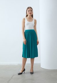 Teal, plissert midikjørt med teksturert overflate, kombinert med en hvit tettsittende tanktop og svarte høye hæler. Gull statement-øreringer.