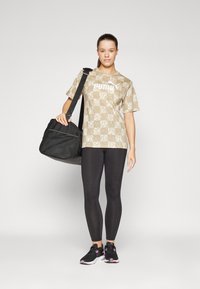 Beige t-skjorte med Puma-mønster, svarte leggings og sneakers. Svart treningsbag med nettpaneler. Modellen står foran en nøytral bakgrunn.
