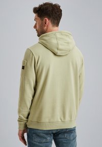 Felpa verde chiaro con cappuccio, polsini e orlo a costine, taschino sul petto e tessuto dal design tassellato. Indossata con jeans blu, in un abbinamento informale.