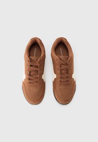 Καφέ αθλητικά παπούτσια από σουέντ με λευκές λεπτομέρειες, στρογγυλή μύτη, επίπεδα κορδόνια και ελαστική σόλα. Ομαλή υφή με δομημένο σχέδιο.