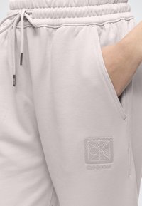 Άτομο που φοράει ανοιχτό γκρι φόρμα Calvin Klein, με το ένα χέρι στην τσέπη, το κορδόνι και το κεντημένο λογότυπο είναι ορατά στο επάνω μέρος του μηρού.