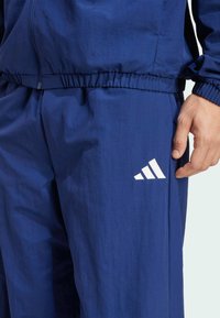 Navy træningsbukser lavet af let materiale med elastisk talje, med et hvidt Adidas-logo på venstre lår.