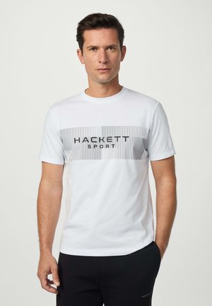 Hombre vestido con una camiseta blanca con rayas verticales grises y el texto "Hackett Sport", acompañado de pantalones negros, de pie frente a un fondo liso.