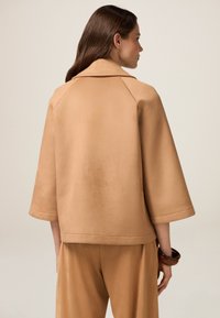 Chaqueta en tono beige, de corte relajado, con cuello y mangas tres cuartos, confeccionada en una tela suave con un ligero brillo. Líneas limpias y detalles mínimos.