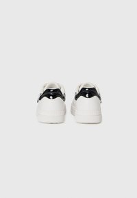 Białe sneakersy o gładkiej fakturze i błyszczącym czarnym akcentem na pięcie. Posiadają zaokrąglone tyły oraz gumową podeszwę. Minimalistyczny design.