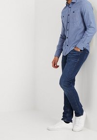 Camicetta blu con bottoni neri, taschino sul petto; abbinata a jeans skinny blu scuro e sneaker bianche. Tessuti lisci.