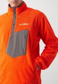 Heldere oranje fleece pullover met een hoge kraag, grijze patser en een grote grijze zak. Zachte textuur door de stof.