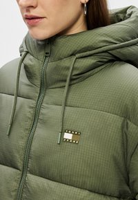 Giacca imbottita verde con colletto alto, dotata di zip integrale, cappuccio con cordino e un patch con logo sul petto. Tessuto testurizzato con lucentezza.