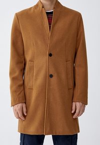 Man in een camelbruine wollen jas met twee zwarte knopen en zijzakken, over een rood geruit overhemd en donkere broeken.