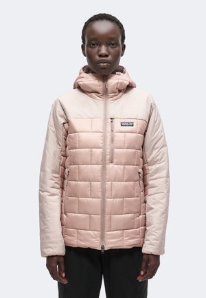Erwachsener in einer hellrosa gesteppte Jacke mit Kapuze, Frontreißverschluss und Reißverschlusstaschen, steht vor einem einfarbigen Hintergrund.