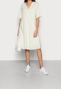 Vihreä lyhythihainen mekko, jossa on V-pääntie, rypytetty vyötärö ja sivutaskut, yhdistettynä valkoisiin tennareihin ja pieneen valkoiseen laukkuun.