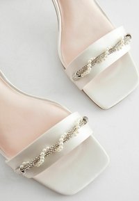 Sandali bianchi in raso con punta quadrata presentano una cintura decorativa con perle e strass, su una soletta morbida rosa.