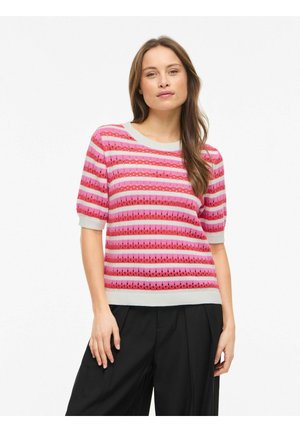 Femme portant un haut tricot à manches courtes rayé rose et rouge avec des manches bouffantes et un pantalon noir à jambes larges, debout sur un fond blanc.