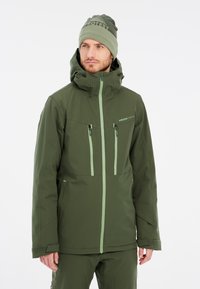 Grüne wasserdichte Jacke mit hohem Kragen, Frontreißverschlüssen und zwei Brusttaschen. Kombiniert mit einer passenden grünen Strickmütze.