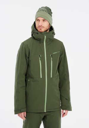 Chaqueta impermeable verde con un alto cuello, cremalleras delanteras y dos bolsillos en el pecho. Combinada con un gorro de punto verde a juego.