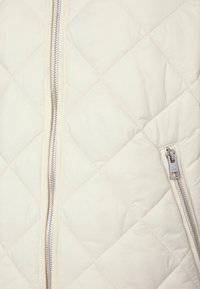 Chaqueta acolchada en color crema con cremalleras plateadas. Presenta costuras con patrón de diamantes y bolsillo lateral con cremallera para mayor funcionalidad.