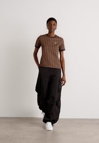 Braun gemustertes T-Shirt mit kurzen Ärmeln, kombiniert mit schwarzen Cargo-Hosen mit seitlichen Taschen und weißen Sneakern.