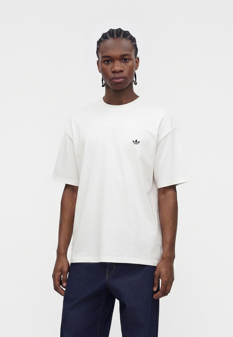 Jeune homme aux cheveux tressés portant un t-shirt blanc Adidas et un pantalon bleu foncé, debout sur un fond clair uni.
