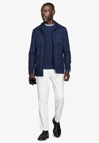 Marineblå knap-frontet jakke med to brystlommer, båret over en marineblå ribbestrikket sweater, parret med hvide bukser og sorte loafers.