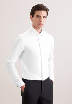 Jeune homme aux cheveux foncés coiffés en arrière, portant une chemise blanche et un pantalon noir, debout les mains dans les poches devant un fond uni.
