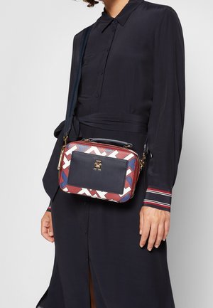 Kvinde iført en sort kjole med knapper og stribede manchetter holder en lille rektangulær crossbody-taske med et geometrisk mønster i rødt, hvidt og blåt.