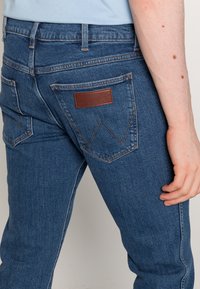 Mörkblå denimjeans med en texturerad yta, med en brun läderpatch på bakfickan och distinkta sömnadsdetaljer.