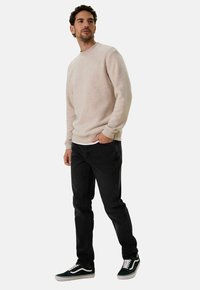 Pull léger beige col rond, jeans noirs et baskets vertes/blanches. Le pull a une texture douce et est ajusté, avec un design classique.