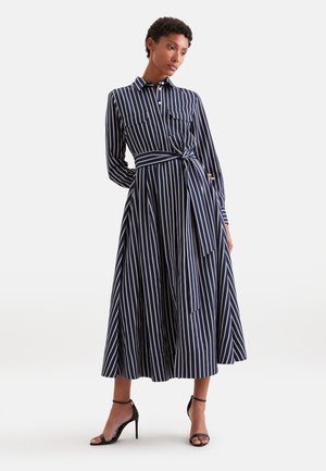 Femme portant une robe midi à manches longues rayée verticalement bleu marine et blanc, avec une ceinture nouée à la taille et des sandales noires à talons hauts.