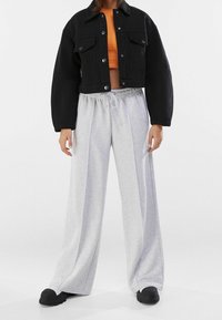 Giacca di jeans nera corta con bottoni argentati, abbinata a pantaloni della tuta grigi chiari a gamba larga e scarpe slip-on nere. È visibile una maglia arancione strutturata.